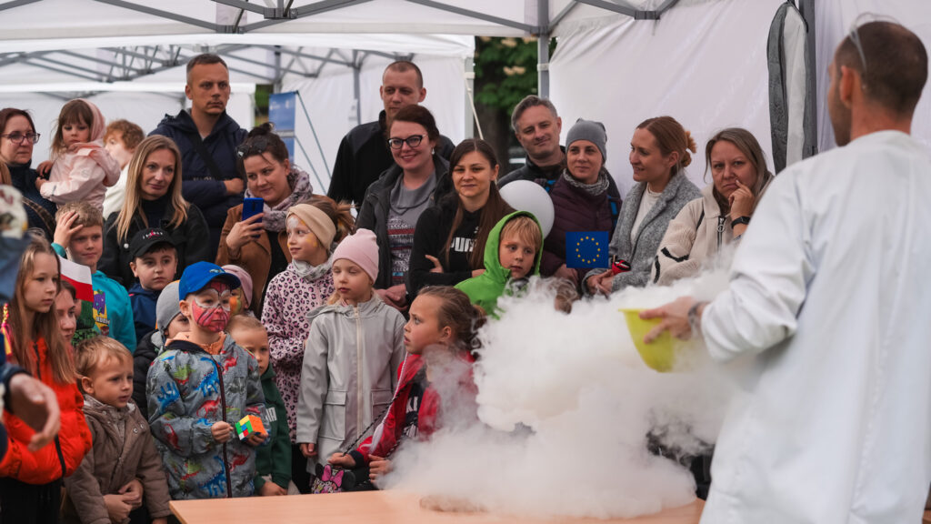 Obrazek przedstawia naukowca w białym fartuchu pokazującego eksperyment z suchym lodem grupie dzieci i dorosłych podczas wydarzenia plenerowego pod namiotem. W tle widać zafascynowaną publiczność.