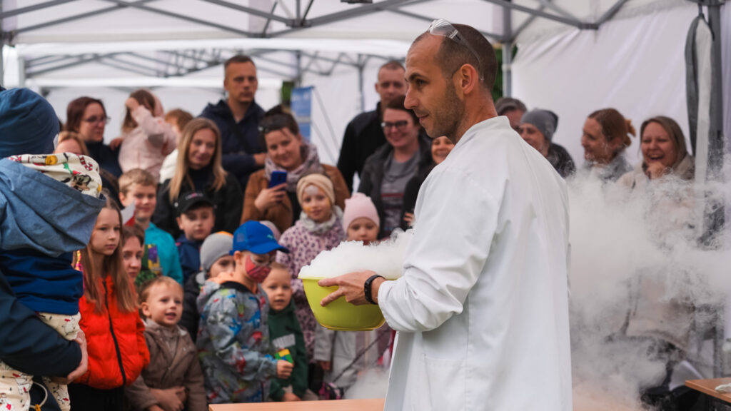 Obrazek przedstawia naukowca w białym fartuchu pokazującego eksperyment z suchym lodem grupie dzieci i dorosłych podczas wydarzenia plenerowego pod namiotem. W tle widać zafascynowaną publiczność.