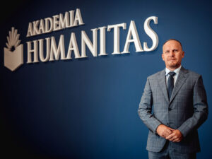 Dr Mariusz Lekston, rektor Akademii Humanitas, pozuje na tle ściany z logo i napisem „Akademia Humanitas”.