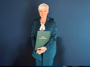 Kobieta trzymająca pracę magisterską pozuje na tle ściany z logo Akademii Humanitas.