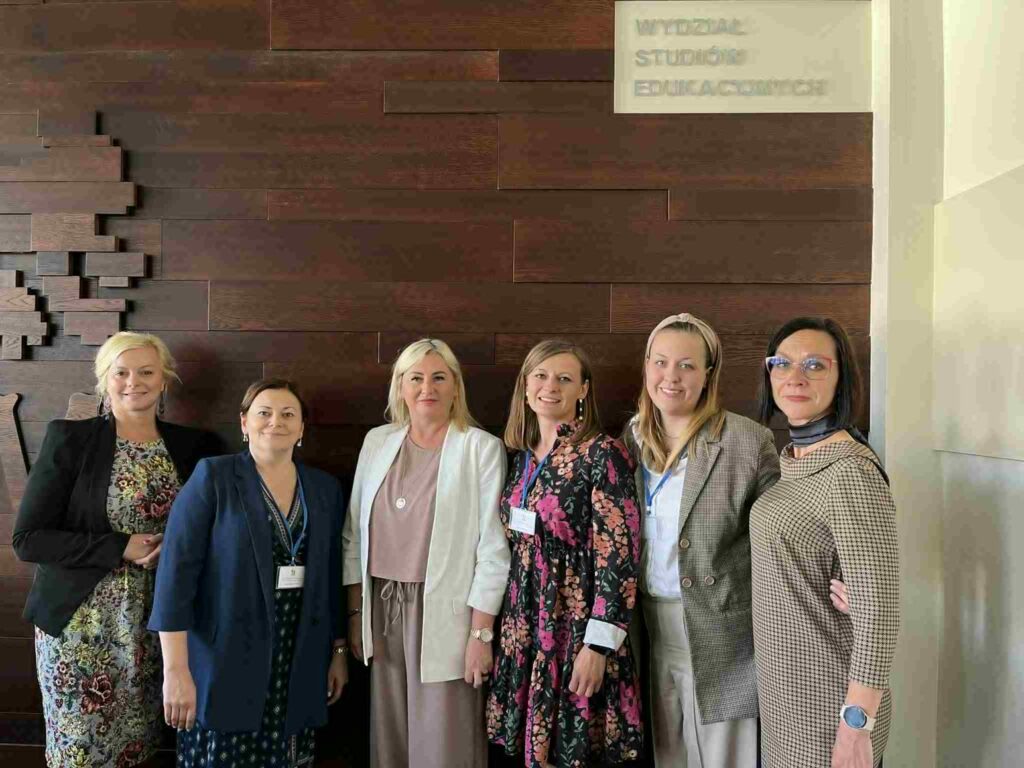 Obrazek przedstawia sześć kobiet uczestniczących w wydarzeniu o charakterze konferencyjnym lub naukowym. Wszystkie osoby są elegancko ubrane i mają identyfikatory, co sugeruje oficjalny charakter spotkania. Tło stanowi dekoracyjna drewniana ściana.