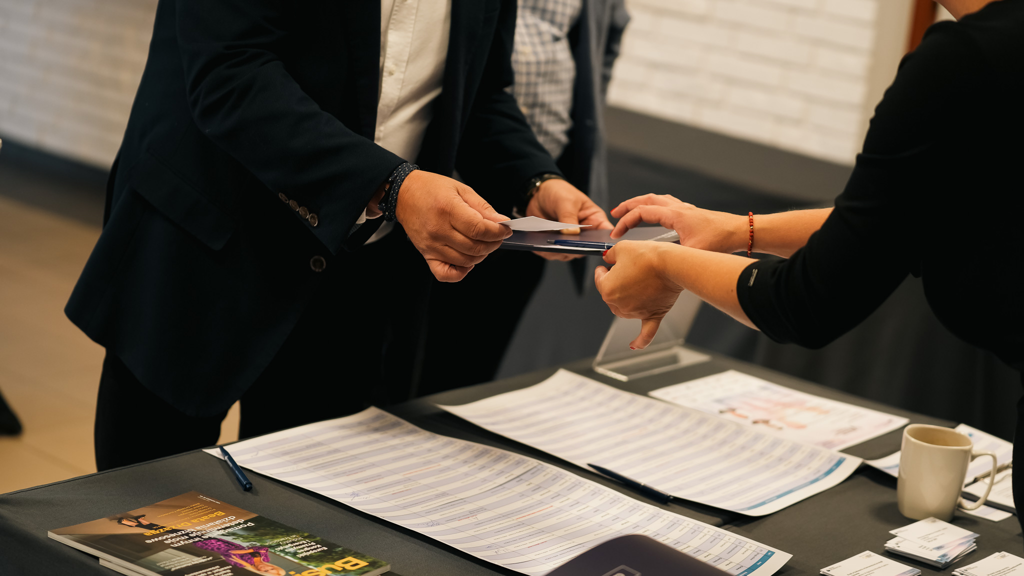 Zbliżenie na moment rejestracji uczestnika wydarzenia. Osoba w garniturze odbiera identyfikator lub materiały od pracownika przy stole rejestracyjnym. Na stole widoczne są listy uczestników, długopisy, wizytówki, czasopismo i filiżanka z kawą