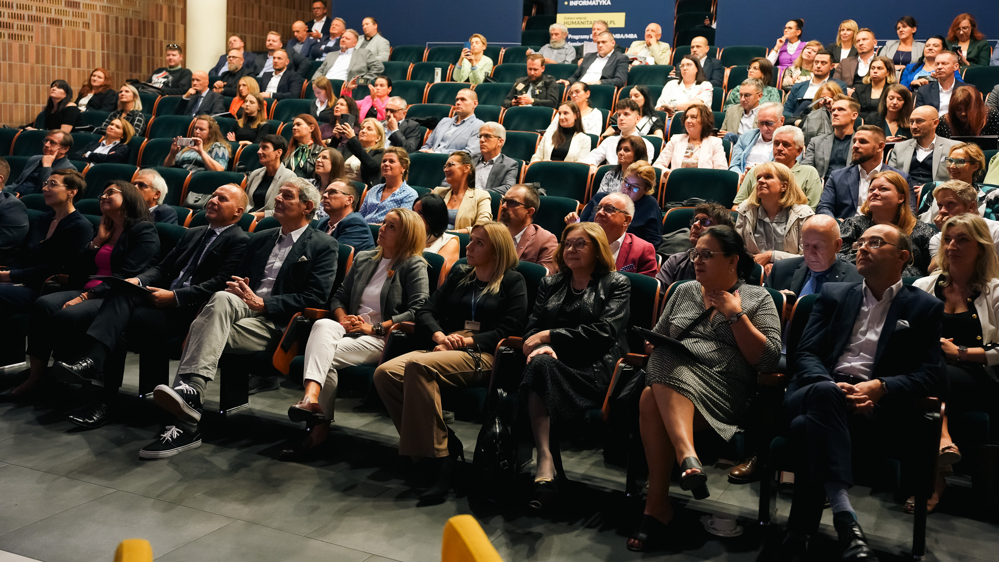 Publiczność zgromadzona w auli podczas konferencji. Uczestnicy siedzą w rzędach zielonych foteli, uważnie słuchając prelekcji. Widać osoby w garniturach, strojach biznesowych i casualowych, część z nich robi notatki lub korzysta z telefonów.