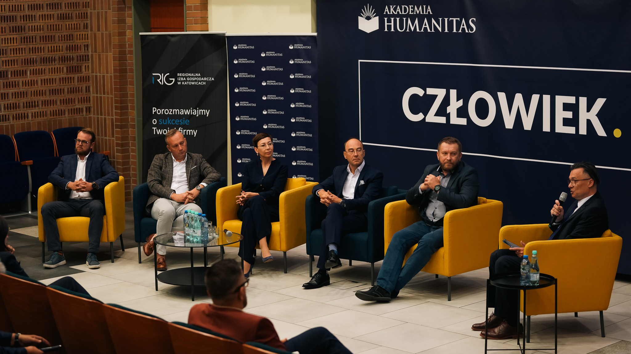 Panel dyskusyjny z udziałem sześciu prelegentów siedzących w fotelach na scenie, prowadzony w Akademii Humanitas. W tle baner z logo Akademii Humanitas i Regionalnej Izby Gospodarczej w Katowicach. Na stolikach stoją butelki z wodą, mikrofony i notatki.