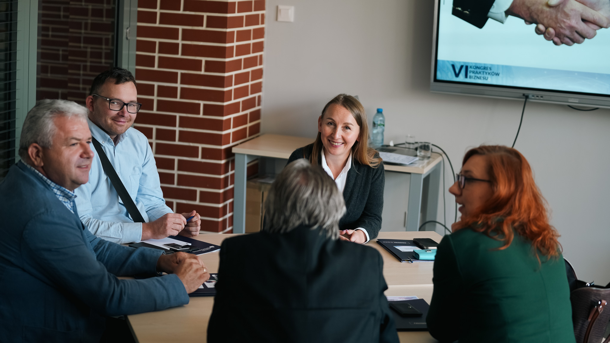 Pięć osób siedzi przy stole w sali konferencyjnej, prowadząc rozmowę i uśmiechając się. Na stole leżą notesy i telefony. W tle widać ekran z fragmentem prezentacji VI Kongresu Praktyków Biznesu oraz butelkę wody i szklanki.