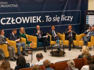 Na zdjęciu widać panel dyskusyjny odbywający się w Akademii Humanitas. Kilku prelegentów – mężczyźni i jedna kobieta – siedzą w kolorowych fotelach na scenie, rozmawiając z publicznością; jeden z nich trzyma mikrofon i prowadzi wypowiedź. Za nimi widnieje duży baner z napisem „CZŁOWIEK. To się liczy”. W pierwszym planie widać część sali z publicznością, która przysłuchuje się dyskusji.