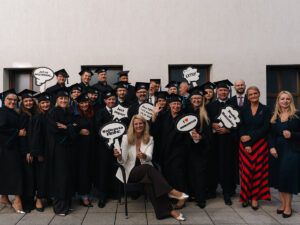 Na zdjęciu widać grupę kilkudziesięciu osób ubranych w czarne togi i birety, stojących razem na tle jasnej ściany budynku. Uśmiechnięci absolwenci pozują do pamiątkowego zdjęcia, trzymając tabliczki z napisami takimi jak „Już po wszystkim!”, „Najlepsza ekipa!”, „Jest super!”, „I ❤️ Humanitas” czy „Studia MBA Humanitas”. Na środku, przed grupą, siedzi kobieta w białej marynarce, również uśmiechnięta i pozująca do zdjęcia.