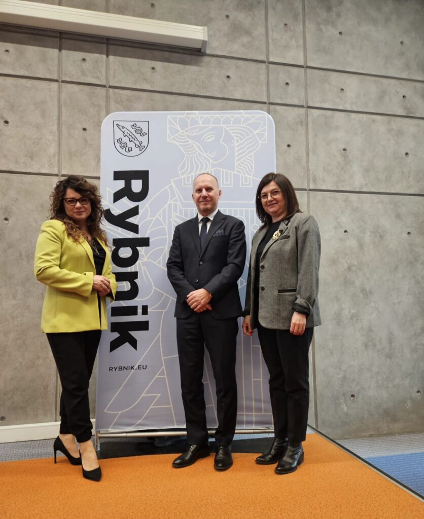 Trzy osoby w eleganckich strojach pozujące przed tablicą z napisem „Rybnik” i herbem miasta.