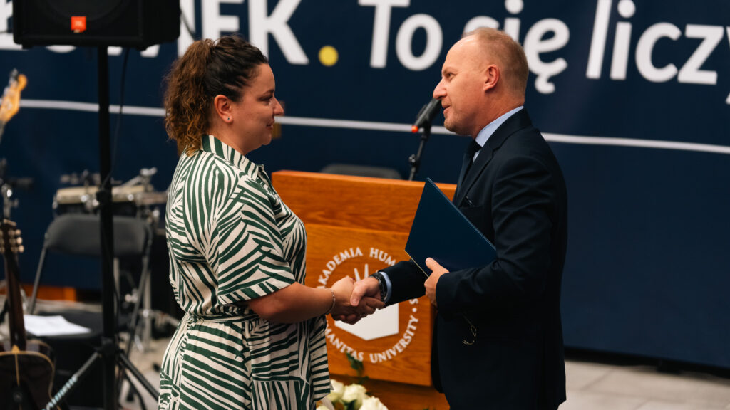Uroczyste wręczenie nagrody koordynatorce UTW w Akademii Humanitas – Rektor AH i koordynatorka UTW podają sobie dłonie przed mównicą uczelni.