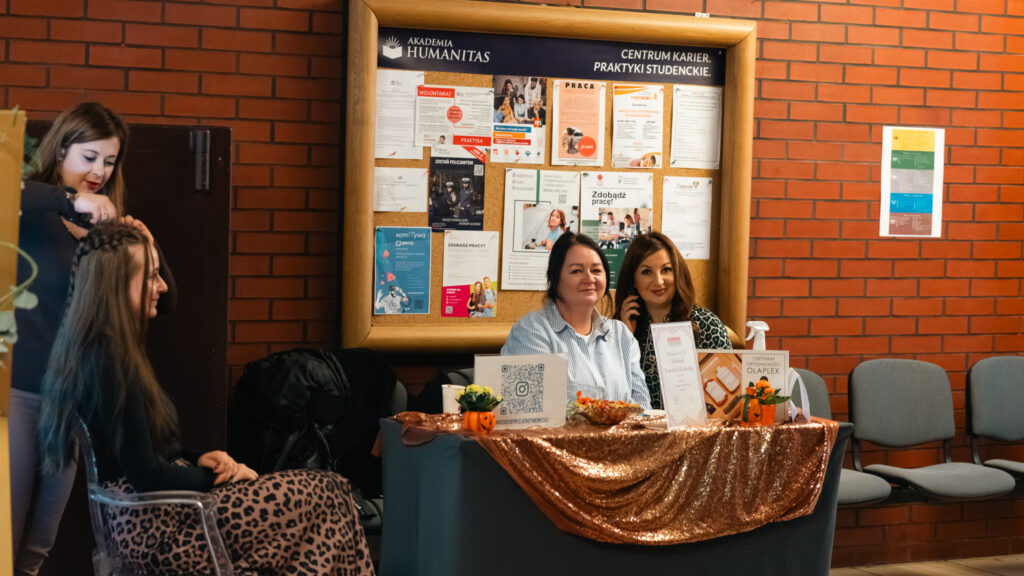 Stoiskosalonu fryzjerskiego, obok którego fryzjerka wykonuje fryzurę uczestniczce wydarzenia.