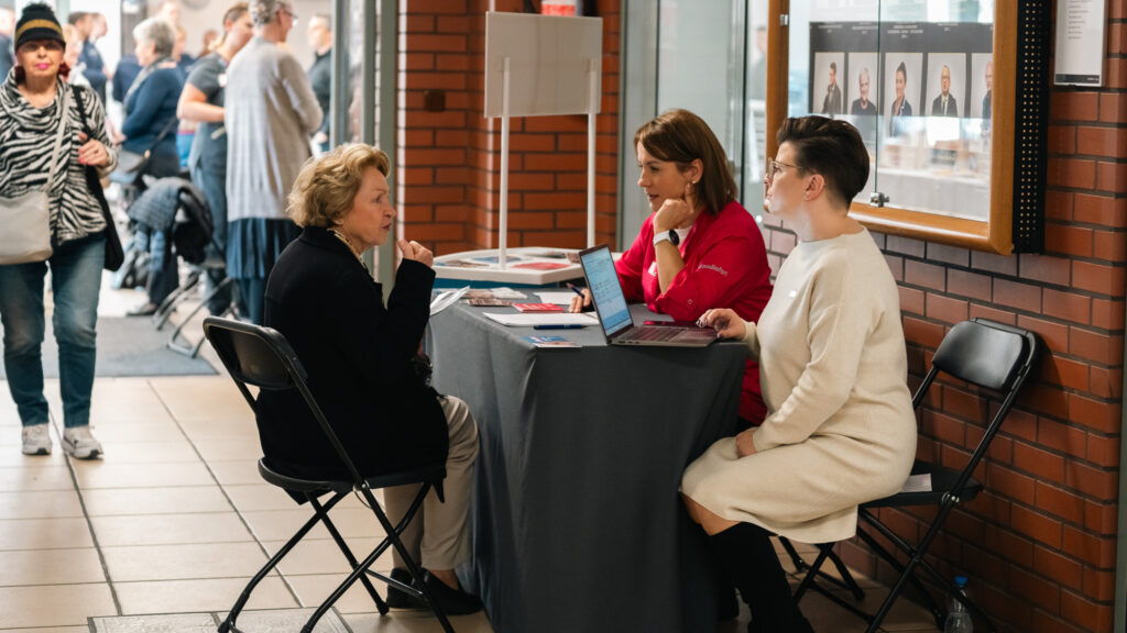 Rozmowa seniorki z dwiema kobietami przy stoliku informacyjnym w Akademii Humanitas podczas wydarzenia dla społeczności lokalnej.