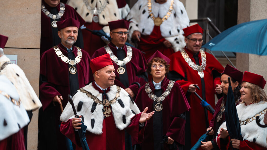 Rektorzy i profesorowie w uroczystych togach i insygniach akademickich idą w orszaku pod parasolami podczas inauguracji roku akademickiego.