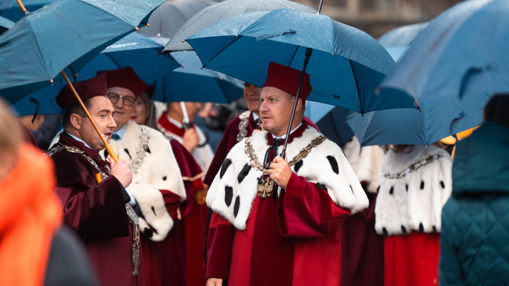 Rektorzy i profesorowie w uroczystych togach i insygniach akademickich idą w orszaku pod parasolami podczas inauguracji roku akademickiego.