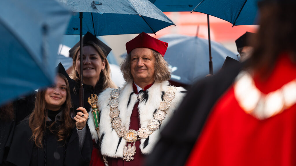 Rektorzy i profesorowie w uroczystych togach i insygniach akademickich idą w orszaku pod parasolami podczas inauguracji roku akademickiego.