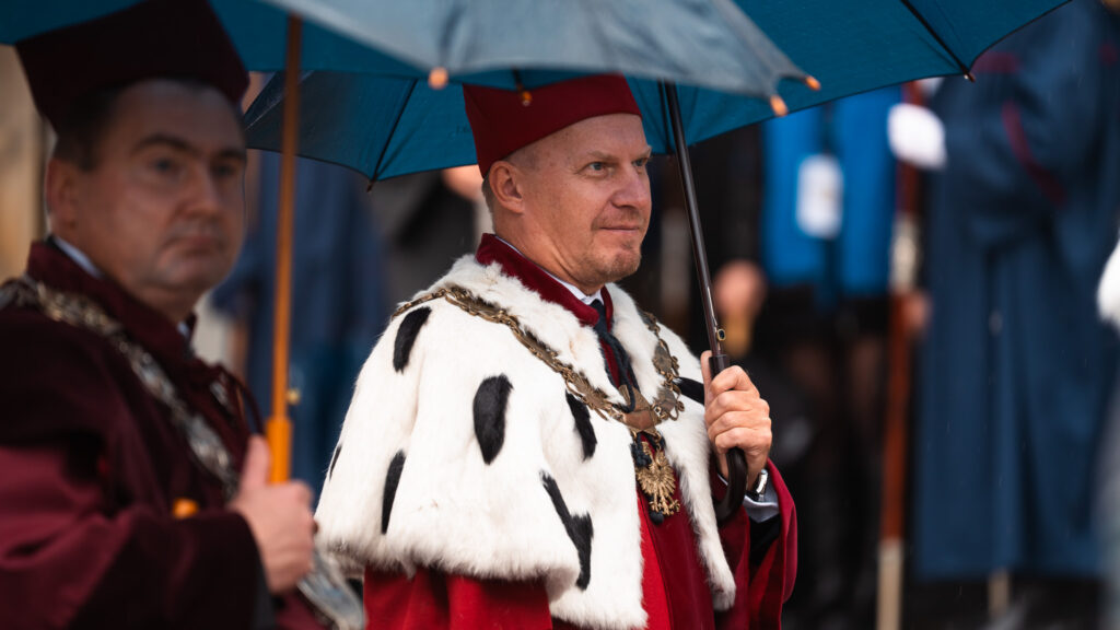 Rektorzy i profesorowie w uroczystych togach i insygniach akademickich idą w orszaku pod parasolami podczas inauguracji roku akademickiego.