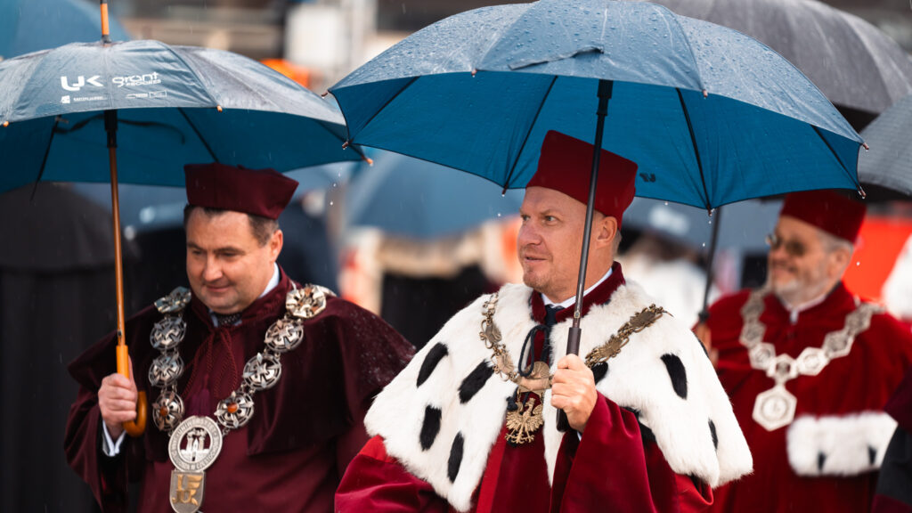 Rektorzy i profesorowie w uroczystych togach i insygniach akademickich idą w orszaku pod parasolami podczas inauguracji roku akademickiego.