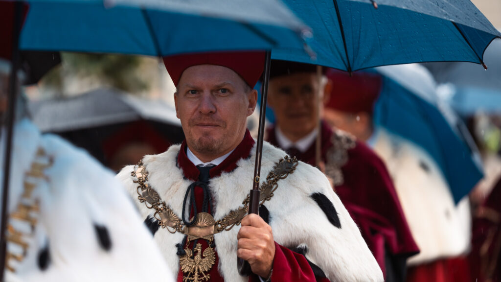 Rektorzy i profesorowie w uroczystych togach i insygniach akademickich idą w orszaku pod parasolami podczas inauguracji roku akademickiego.