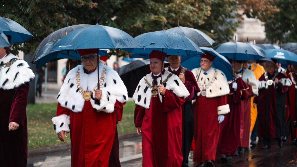 Rektorzy i profesorowie w uroczystych togach i insygniach akademickich idą w orszaku pod parasolami podczas inauguracji roku akademickiego.