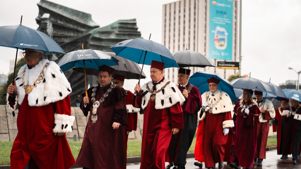 Rektorzy i profesorowie w uroczystych togach i insygniach akademickich idą w orszaku pod parasolami podczas inauguracji roku akademickiego.