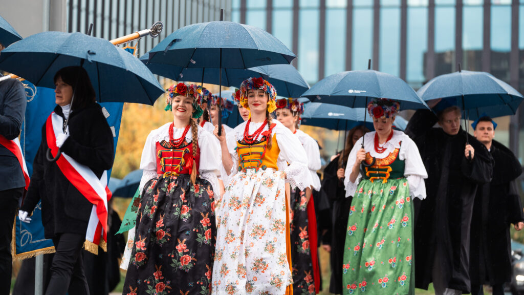 Kobiety w tradycyjnych strojach ludowych idą z parasolami podczas akademickiej inauguracji.