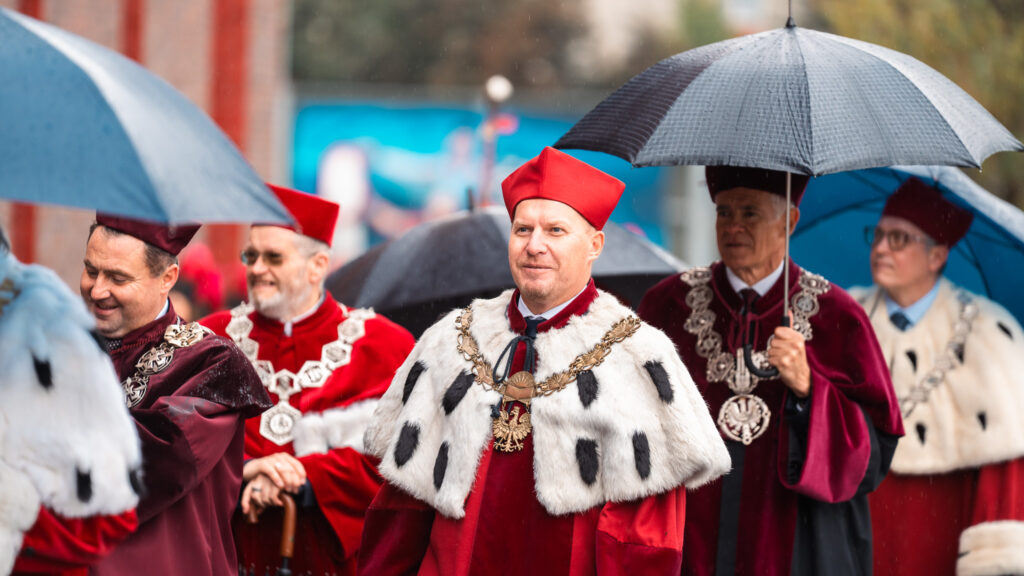 Rektorzy i profesorowie w uroczystych togach i insygniach akademickich idą w orszaku pod parasolami podczas inauguracji roku akademickiego.