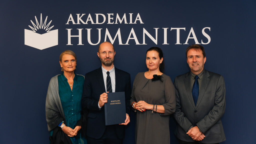 Cztery osoby w eleganckich strojach pozują na tle granatowej ściany z logo Akademii Humanitas; jedna z nich trzyma teczkę z napisem „Rozprawa doktorska”.