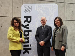 Trzy osoby w eleganckich strojach pozujące przed tablicą z napisem „Rybnik” i herbem miasta.