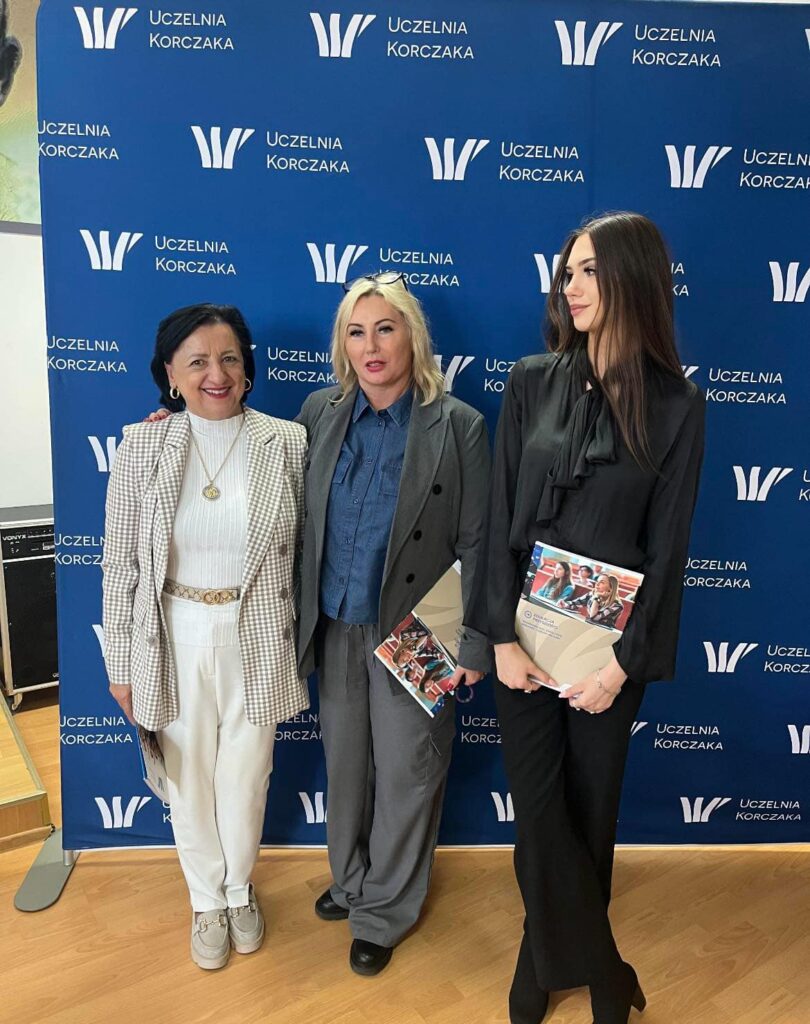 Trzy kobiety pozujące na tle ścianki z logo Uczelni Korczaka, trzymające materiały informacyjne uczelni.
