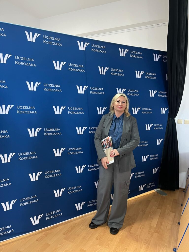 Kobieta na tle ścianki z logo Uczelni Korczaka, trzymające materiały informacyjne uczelni.