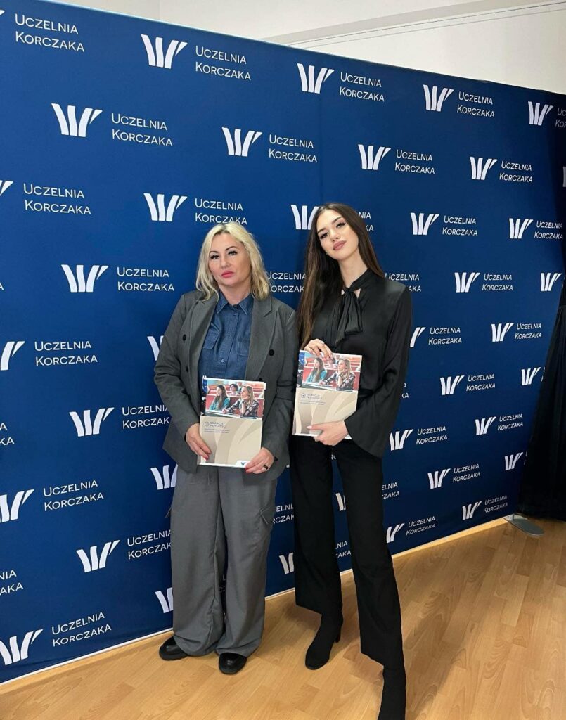 Dwie kobiety pozujące na tle ścianki z logo Uczelni Korczaka, trzymające materiały informacyjne uczelni.