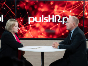 Wywiad w studiu PulsHR.pl – kobieta - redaktorka - i mężczyzna - rektor Akademii Humanitas - rozmawiają przy stoliku na tle czerwonej scenografii z logo portalu.