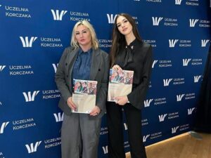 Dwie kobiety pozujące na tle ścianki z logo Uczelni Korczaka, trzymające materiały informacyjne uczelni.