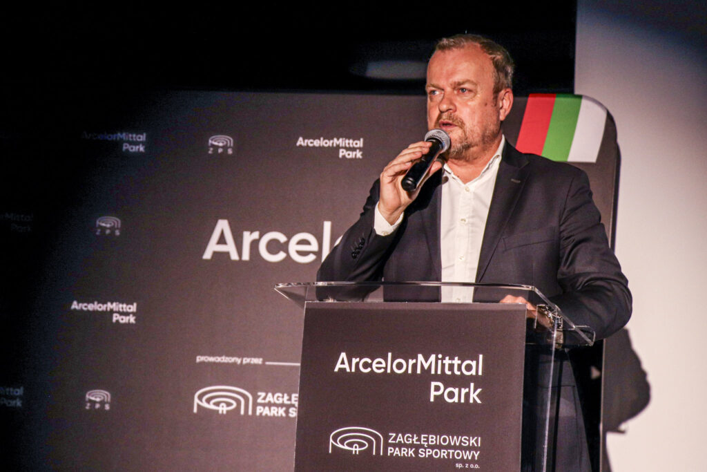 Mężczyzna w garniturze przemawia do mikrofonu, stojąc za mównicą z logotypem ArcelorMittal Park podczas oficjalnego wydarzenia.