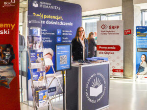 Stoisko Akademii Humanitas z materiałami informacyjnymi podczas wydarzenia – za ladą stoi przedstawicielka uczelni, a wokół znajdują się roll-upy promocyjne i stojak z ulotkami.
