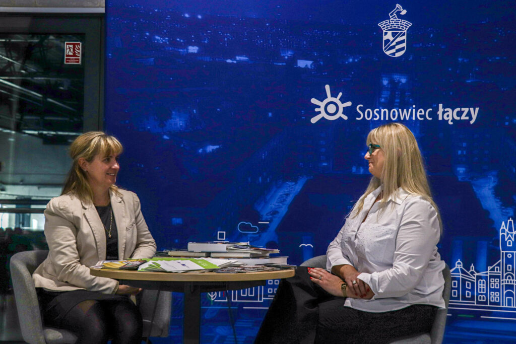 Dwie kobiety siedzą przy okrągłym stoliku i rozmawiają; na stole leżą książki i ulotki. W tle duża niebieska grafika z panoramą miasta oraz napisem ‘Sosnowiec łączy’ i herbem miasta.