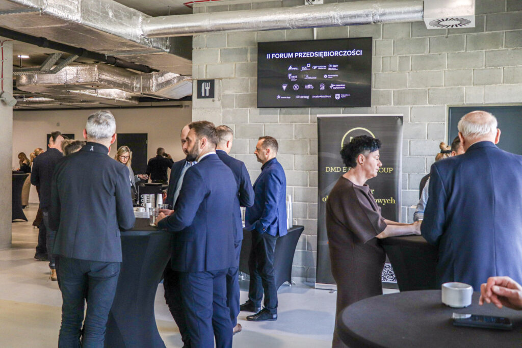 Grupa elegancko ubranych osób rozmawia przy wysokich stolikach podczas wydarzenia II Forum Przedsiębiorczości, odbywającego się w nowoczesnej sali z betonowymi ścianami i widocznymi instalacjami sufitowymi.
