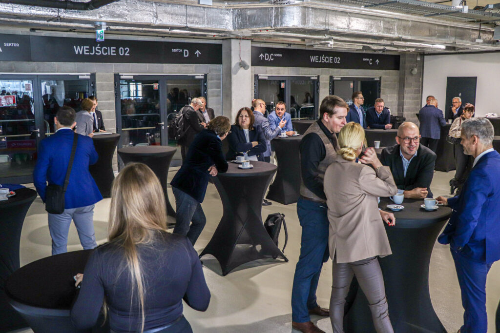 Grupa elegancko ubranych osób rozmawia przy wysokich stolikach podczas wydarzenia II Forum Przedsiębiorczości, odbywającego się w nowoczesnej sali z betonowymi ścianami i widocznymi instalacjami sufitowymi.