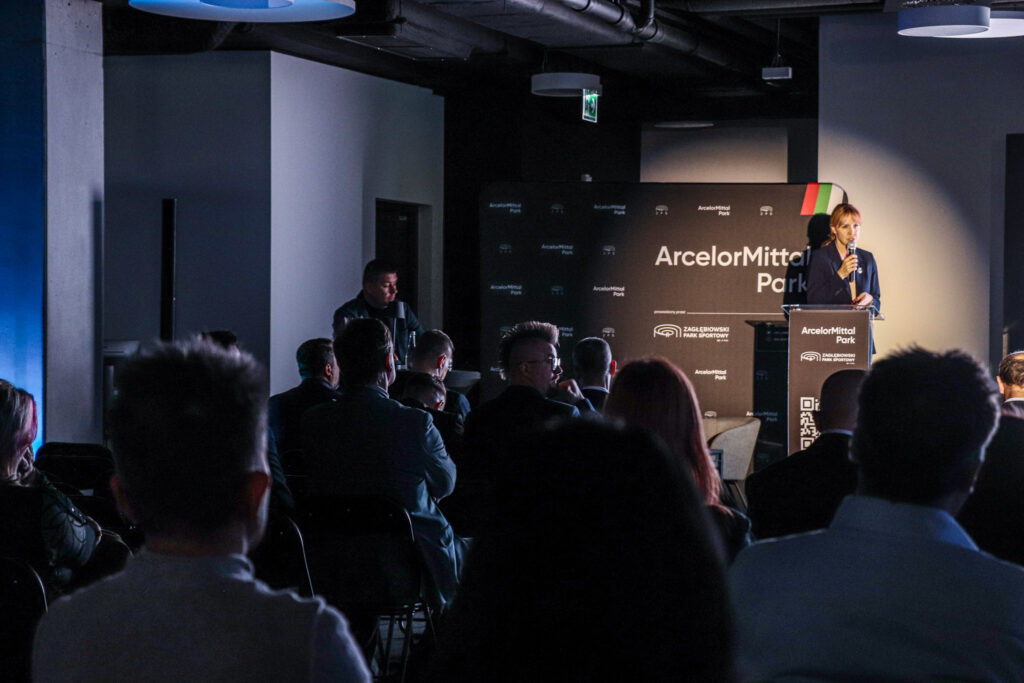 Prelegentka stojąca na scenie przemawia do publiczności podczas wydarzenia ArcelorMittal Park; widownia siedzi w półmroku, a scena jest oświetlona punktowo.