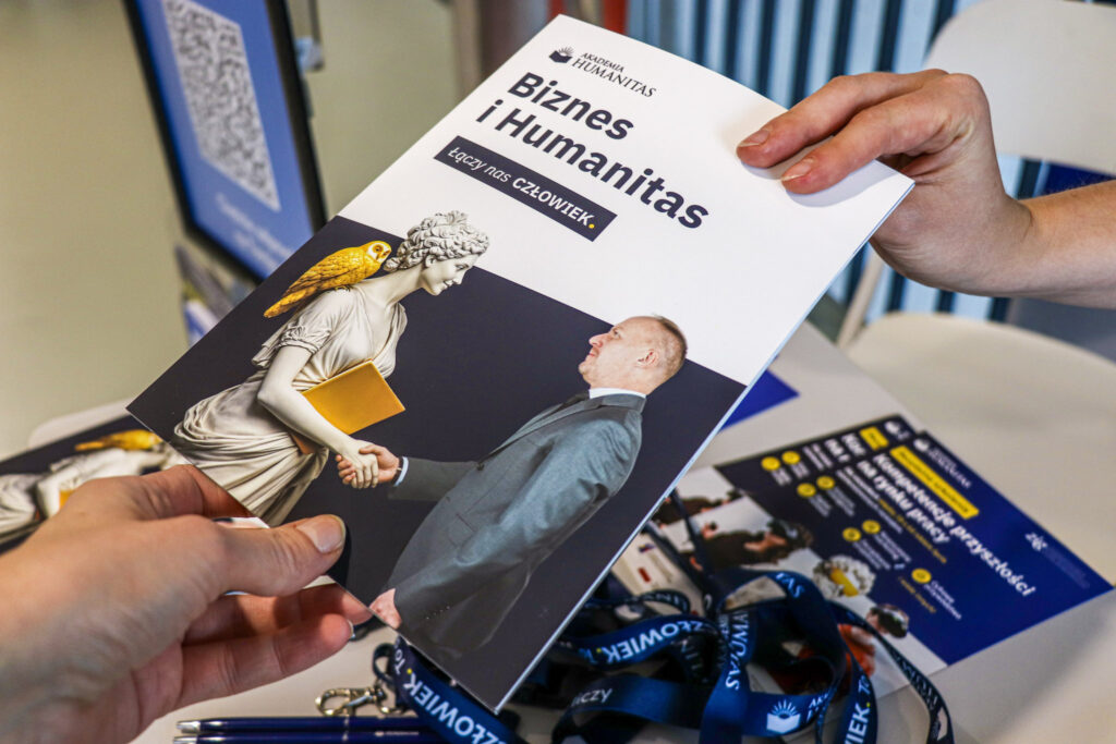 Ujęcie zbliżone na ulotkę „Biznes i Humanitas”, trzymaną przez dwie osoby. Na okładce widoczna jest ilustracja przedstawiająca uścisk dłoni między klasyczną rzeźbą a elegancko ubranym mężczyzną. W tle na stole leżą smycze i inne materiały informacyjne.