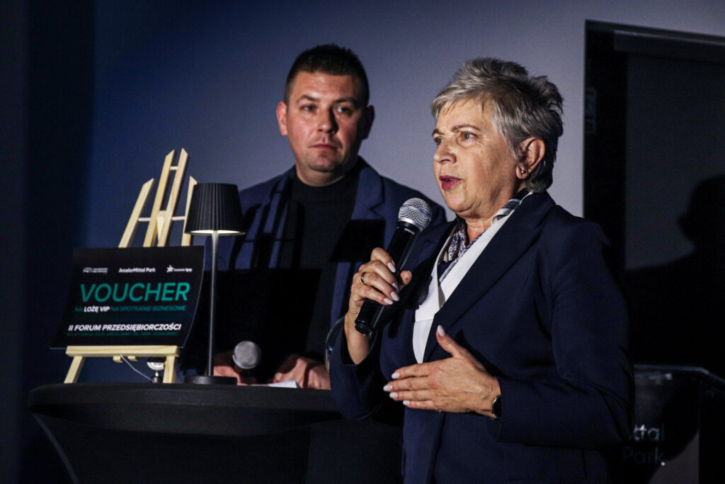 Kobieta w granatowej marynarce przemawia do mikrofonu podczas wydarzenia biznesowego, stojąc obok mężczyzny przy wysokim stoliku z voucherem na spotkanie biznesowe.