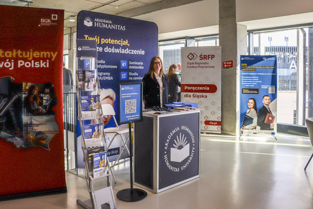 Stoisko Akademii Humanitas z materiałami informacyjnymi podczas wydarzenia – za ladą stoi przedstawicielka uczelni, a wokół znajdują się roll-upy promocyjne i stojak z ulotkami.