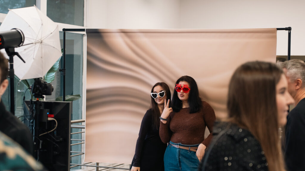 Dwie młode kobiety pozują do zdjęcia na tle jasnego, falowanego tła fotograficznego, mając na sobie duże, kolorowe okulary w zabawnym kształcie. Jedna z nich pokazuje znak pokoju. Wokół nich widoczne są elementy studia fotograficznego oraz inni uczestnicy wydarzenia.