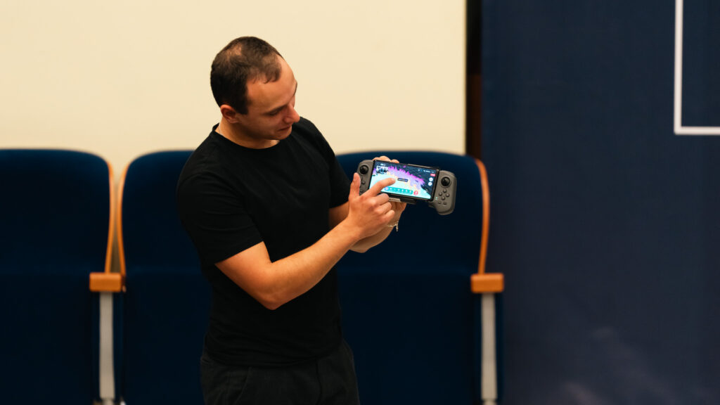 Mężczyzna w czarnym T-shircie prezentuje na sali wykładowej przenośną konsolę do sterowania robotem trzymając ją w dłoniach i wskazując palcem na ekran.