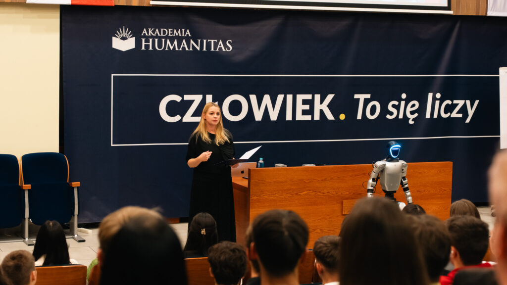 Kobieta stojąca za mównicą podczas wykładu w auli Akademii Humanitas, obok humanoidalny robot; na tle widoczny duży baner z napisem ‘Człowiek. To się liczy’.