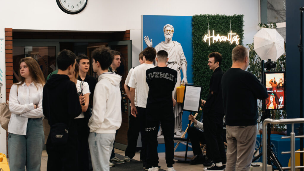 Grupa studentów stojących w holu obok dekoracji z białą rzeźbą w okularach i neonowym napisem ‘#Humanitas’; część osób rozmawia, inni czekają przy stanowisku fotograficznym z lampą i ekranem.