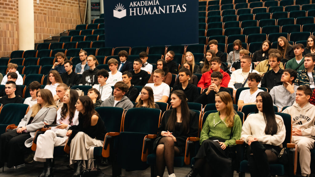 Grupa młodych osób siedzi w audytorium Akademii Humanitas, uważnie słuchając prelekcji; widoczne rzędy zielonych foteli oraz tło z logotypem uczelni.