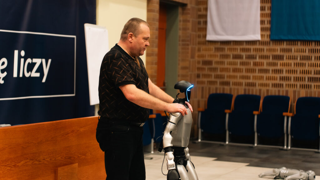 Mężczyzna prezentuje humanoidalnego robota podczas spotkania w auli, trzymając go za ramiona. Robot ma świecący na niebiesko panel na głowie, a w tle widać rzędy krzeseł i fragment sceny.