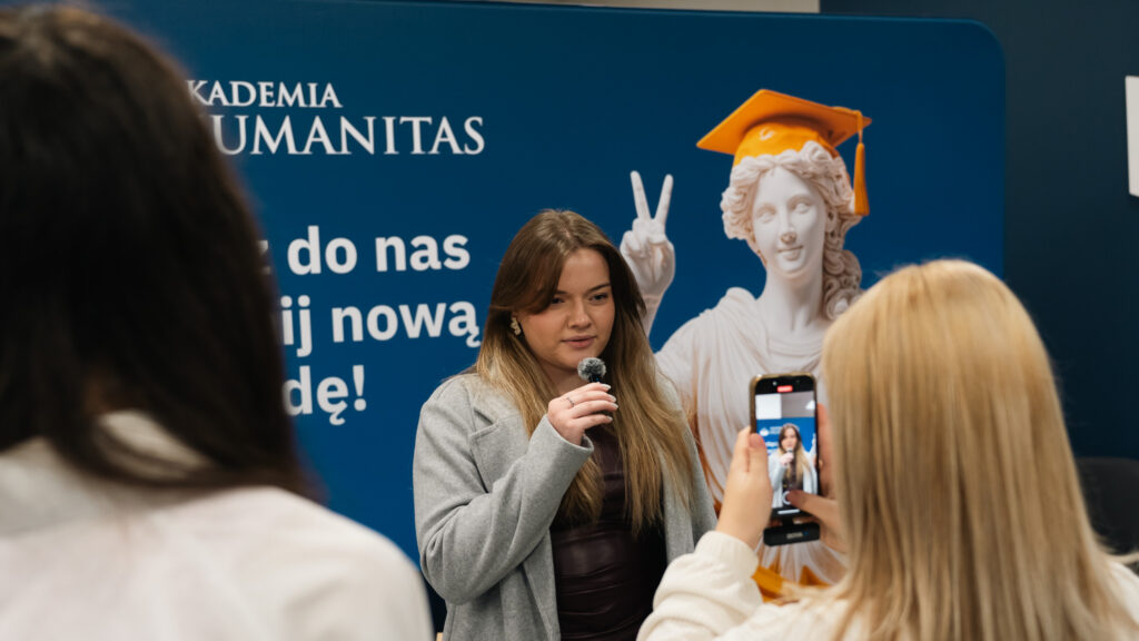 Młoda kobieta trzyma mikrofon i udziela wypowiedzi, podczas gdy inna osoba nagrywa ją telefonem. W tle widoczna jest niebieska ścianka Akademii Humanitas z napisem oraz rzeźba w birecie wykonująca gest pokoju.