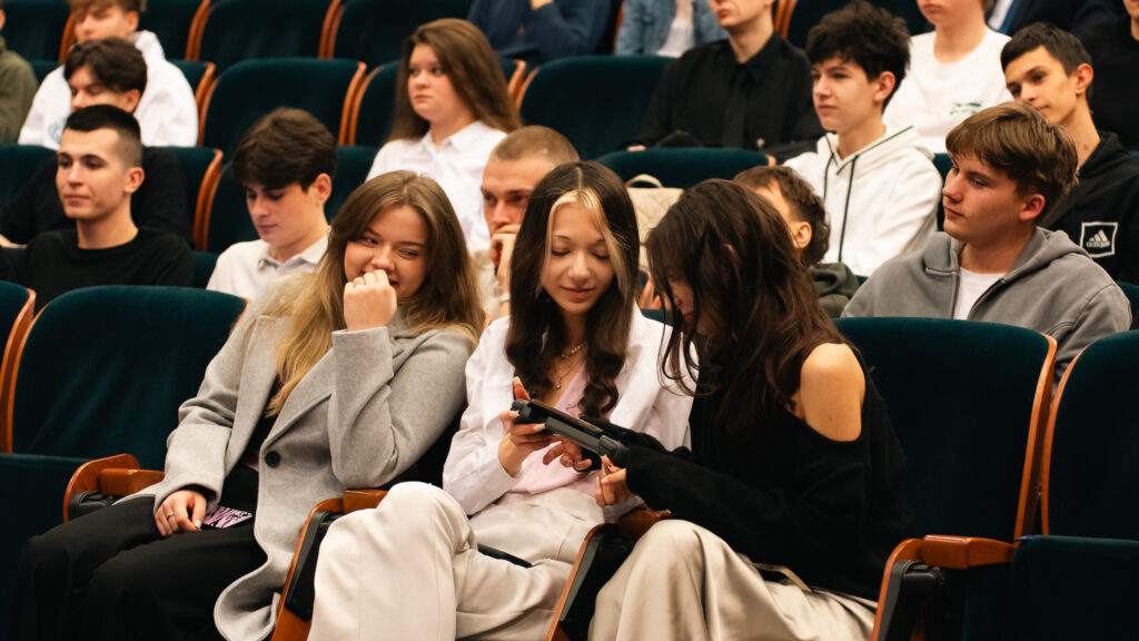 Trzy młode kobiety siedzą w audytorium, uśmiechają się, wokół nich pozostali uczestnicy wydarzenia.