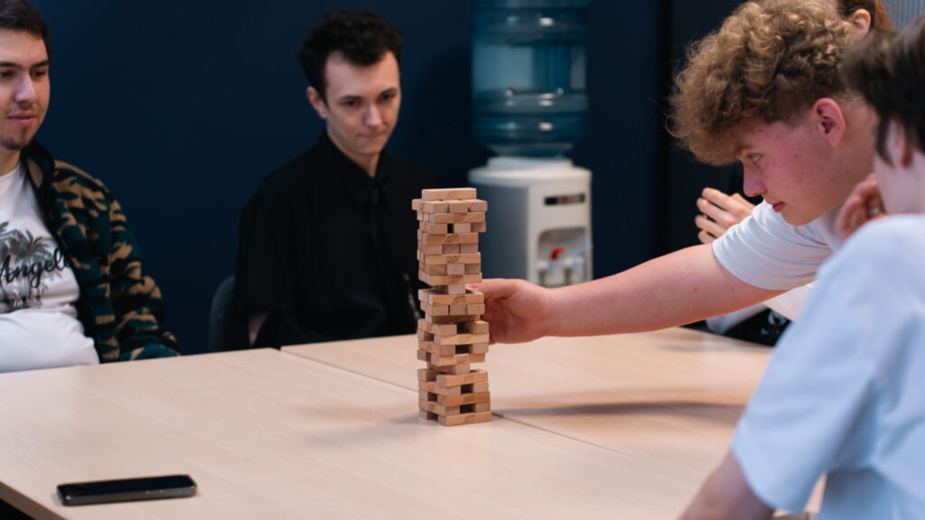 Grupa młodych osób siedzi przy stole i gra w Jengę; jeden z chłopców sięga po klocek, a pozostali uczestnicy obserwują grę w skupieniu.